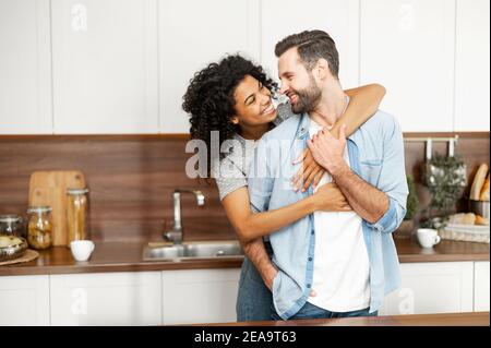 Primo piano di una felice coppia interracial in piedi in cucina, felici proprietari romantici di un nuovo appartamento sorridente e guardandosi l'un l'altro, giovane africano Foto Stock