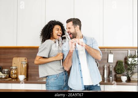 Felice coppia interracial danzare in cucina, cantando mentre cucinando la prima colazione o la cena, giovane donna afroamericana e un bell'uomo sorridente Foto Stock