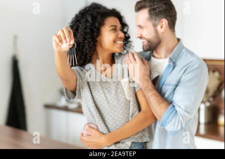 Felice giovane coppia sposata interracial che tiene un anello chiave in mano, abbracciando e guardandosi l'un l'altro con amore, in piedi nella cucina moderna di Foto Stock