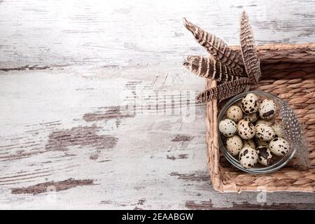 Quail uova in una ciotola di vetro e piume in giacinto d'acqua cestello di stoccaggio rettangolare su tavolo di legno. Spazio di copia, modello, vista dall'alto Foto Stock