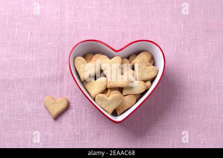 Deliziosi biscotti fatti in casa cuori in una ciotola a forma di cuore rosso per San Valentino su uno sfondo di tessuto rosa, vista dall'alto Foto Stock