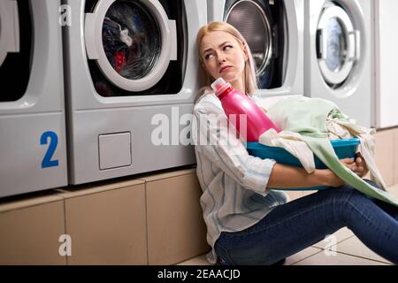 Annoiata donna che tiene vestiti sporchi e detersivo bottiglia rosa in bacino, si siede sul pavimento appoggiandosi sulla lavatrice, sedersi depresso da solo, in lavaggio ho Foto Stock