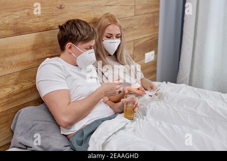 Coppia in maschere mediche prendendo farmaci pillole vitamine mentre si siede a letto che soffrono di COVID-19, sono messi in quarantena a casa insieme. Soggiorno ho Foto Stock