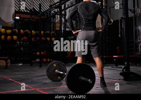 vista posteriore su sportivo in piedi vicino al barbell prendendo pausa, riposo. maschio in outfit sportivo in moderna palestra Foto Stock