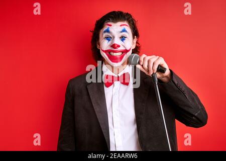 mago con microfono isolato su sfondo rosso, gioioso trucco magico uomo o mistero uomo in vestito nero parlando, sorridente Foto Stock