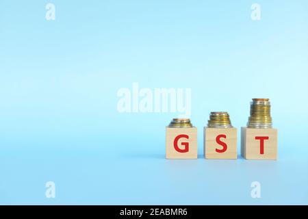 Lettere GST su blocchi di legno su sfondo blu crescente pila di monete. Aumento del concetto di imposta sui beni e sui servizi. Foto Stock