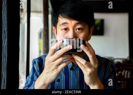 Giovane uomo cinese adulto in costume tradizionale che beve una tazza di tè caldo nel ristorante cinese. Un ragazzo asiatico attento ha un sorso di bevande rinfrescanti vicino alla finestra del caffè. Foto Stock