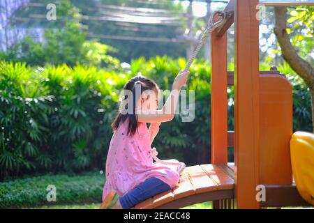 Una giovane ragazza asiatica carina che gioca una scala di legno con la corda di arrampicata in un parco giochi pubblico con erba verde e albero e una giornata di sole. Foto Stock