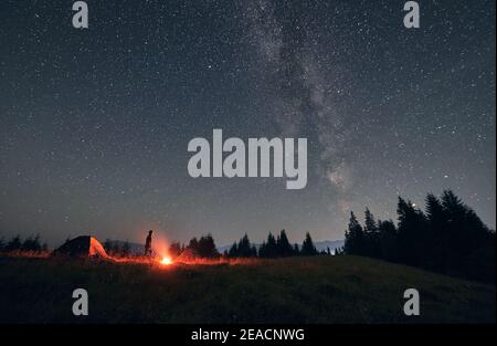 Escursionista maschile in piedi vicino al falò e tenda sotto il bellissimo cielo notturno con stelle. Magnifica vista del cielo stellato blu e della Via Lattea sotto la collina erbosa. Concetto di viaggio, escursioni e campeggio notturno. Foto Stock