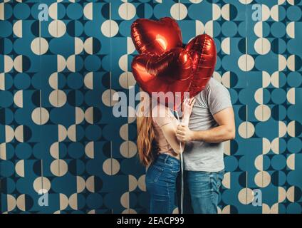 Baciare la coppia caucasica si nascondono dietro un po 'di aria cuore affilato palloncini su una parete blu dello studio Foto Stock