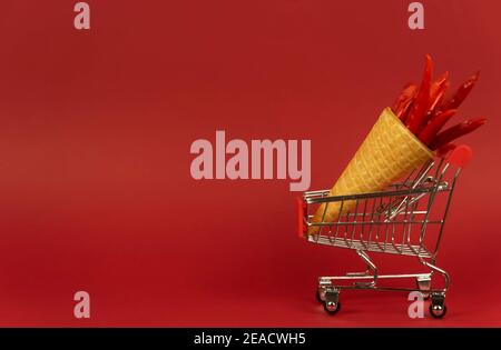 Un cono di cialda pieno di peperoncini rossi caldi giace in un carrello per lo shopping su sfondo rosso Foto Stock