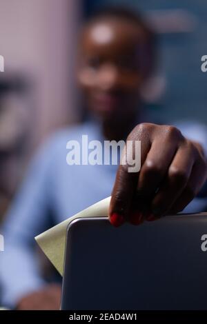 Primo piano di una donna d'affari africana che usa note appiccicose sul computer portatile che lavora in remoto dall'ufficio domestico seduto alla scrivania. Freelance nero rispettando la scadenza di studio a tarda notte. Foto Stock