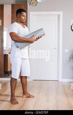 Ripresa ad angolo basso di un muscoloso sportivo afroamericano che rotola il tappeto yoga dopo aver finito di allenarsi. Foto Stock