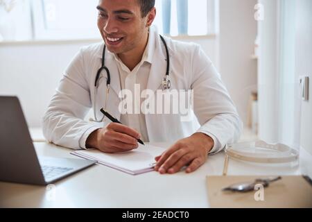 Bel medico professionista in accappatoio bianco seduto al tavolo mentre si guarda allo schermo del computer portatile Foto Stock