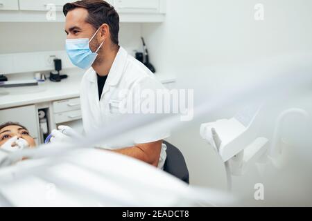 Dentista maschile che parla con il suo assistente mentre esamina i denti della paziente femminile in clinica. Dentista in maschera protettiva per il viso durante il trattamento di un paziente in Foto Stock