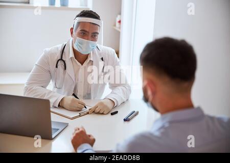 Bel dottore maschile in bianco accappatoio seduto al tavolo davanti al paziente Foto Stock