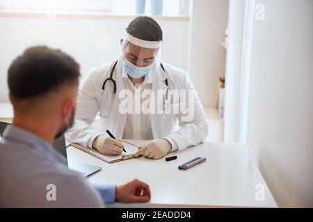 Bel dottore maschio che scrive qualcosa su carta bianca mentre ascolta il suo paziente in camera al chiuso Foto Stock