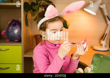 Una ragazza nelle orecchie di un coniglietto pasquale dipinge le uova con una penna di feltro-punta nell'interno domestico. Artigianato, preparazione per una festa religiosa, un vassoio con e. Foto Stock