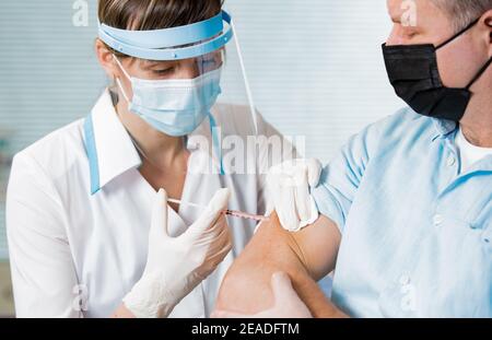 Medico femminile con maschera chirurgica e in guanti che danno iniezione di vaccino all'uomo in ospedale. Vaccinazione durante la pandemia COVID-19 Foto Stock