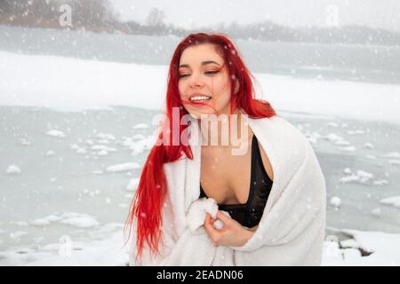 Ritratto invernale innevato di una donna rossa all'aperto in costume da bagno, avvolto in un asciugamano al lago di nuoto in acqua aperta, terapia fredda e indurimento sulla neve Foto Stock