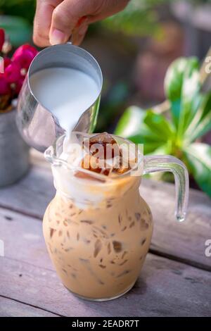 Versare il latte in un bicchiere con caffè latte e ghiacciato su un tavolo di legno. Foto Stock