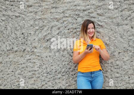 Sorridente donna caucasica millenaria che tiene un telefono cellulare e si appoggia contro un muro di pietra, scorrendo lo schermo e guardandola a sinistra Foto Stock
