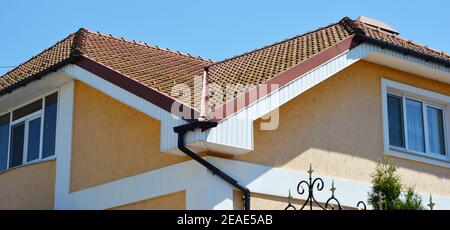 Un primo piano su una valle del tetto in piastrelle di argilla rossa, fascia, box end, soffit, bareboard e sistema di gocciolatoio con un tubo discendente di una casa dipinta. Foto Stock