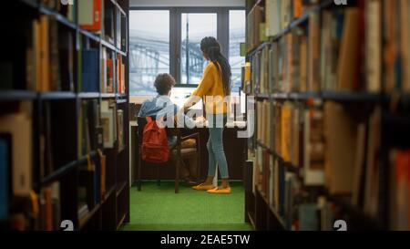 Biblioteca universitaria: Il ragazzo usa il personal computer alla sua scrivania, parla con ragazza Classmate che spiega, lo aiuta con assegnazione di classe. Studenti concentrati Foto Stock