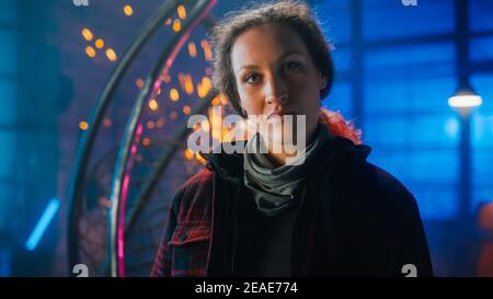 Giovane bella Empowerering Donna con l'orecchio Piercing sorride delicatamente alla macchina fotografica. Autentico Fabricator che indossa abiti da lavoro in un laboratorio di metallo. Scintille Foto Stock