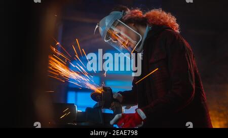 La talentuosa Female Fabricator utilizza una smerigliatrice angolare per lucidare un oggetto metallico circolare. L'anello è afferrato in una morsa da banco. Bellissimo Tomboy Engineer Foto Stock