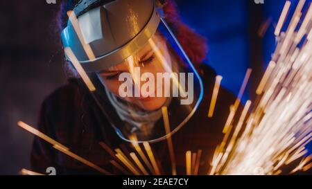 Primo piano Ritratto di bella Femminile Fabricator in maschera di sicurezza. Sta macinando un oggetto metallico e Flying Hot Sparks riflettere sulla maschera. Potenziamento Foto Stock