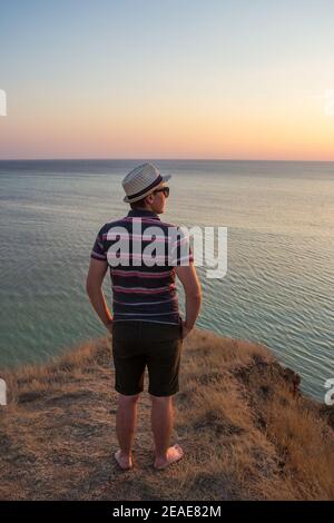 Un ragazzo in shorts e cappello di paglia in piedi su un scogliera sul mare Foto Stock