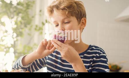 In cucina: Adorabile Boy mangia cremoso Cupcake con glassatura e spolverato Funfetti. Carino affamato Sweet Tooth Child morde in Muffin con zuccherato Foto Stock