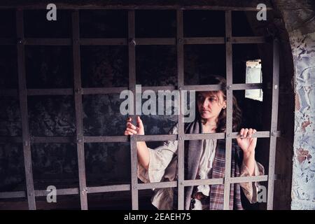 Una donna guarda attraverso le sbarre di una vecchia cella di prigione. Una giovane donna in un abito d'epoca del 18-19 secolo. Foto Stock