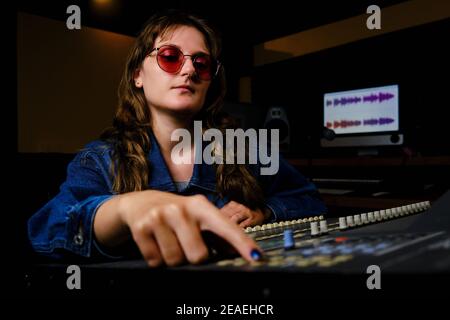 Donna ingegnere del suono al mixer in studio di musica. Donna in occhiali da sole e jeans dietro console di mixaggio in studio di registrazione. Foto Stock