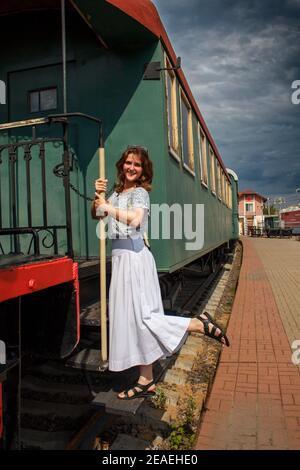 Donna in piedi sul bordo di corsa dell'automobile dell'era sovietica. Tenere il corrimano del treno in partenza. Piattaforma vuota e cielo coperto. Auto d'epoca o Foto Stock