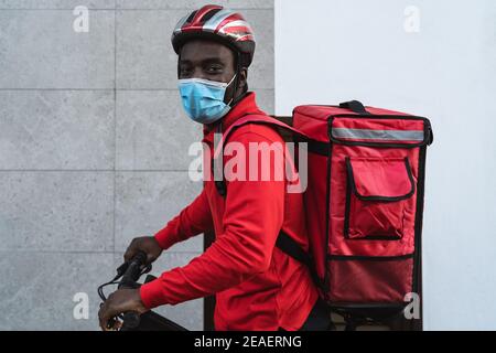 Pilota africano che consegna il pasto ai clienti con bicicletta elettrica mentre indossa la maschera facciale durante l'epidemia di virus corona Foto Stock