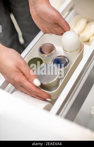 Mani donna che tengono contenitore di plastica con creme e collocano nel cassetto della stanza da bagno. Foto Stock