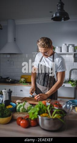 Bell'uomo in camicia bianca e grembiule sta facendo un sano pasto biologico di insalata in una moderna cucina Sunny. Hipster Man in cucina bicchieri. Pulizia naturale Foto Stock