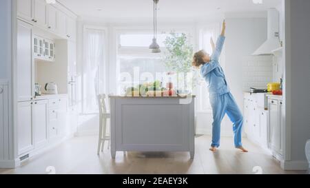 Energico giovane uomo divertente con lunghi capelli che ballano in cucina mentre indossa le Pajama blu. Area cucina moderna di colore bianco luminoso con verde sano Foto Stock