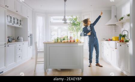 Energico giovane uomo divertente con lunghi capelli che ballano in cucina mentre indossa una tuta blu. Area cucina moderna di colore bianco luminoso con verde sano Foto Stock