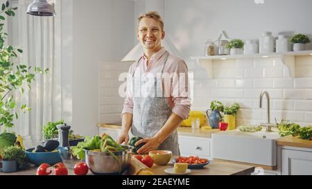 Bell'uomo in camicia rosa e grembiule sta facendo un sano pasto biologico insalata in una moderna cucina Sunny. Hipster Man in occhiali sorride alla macchina fotografica Foto Stock