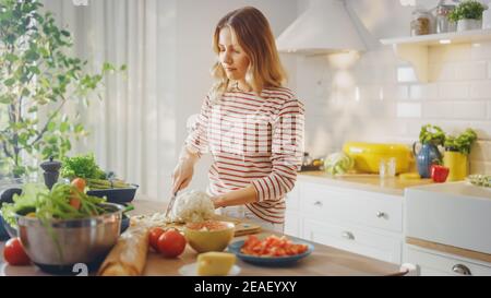 Giovane femmina in ponticello a strisce sta facendo una sana insalata biologica in una moderna cucina soleggiato. La donna autentica sta tagliando un cavolfiore con un coltello Foto Stock