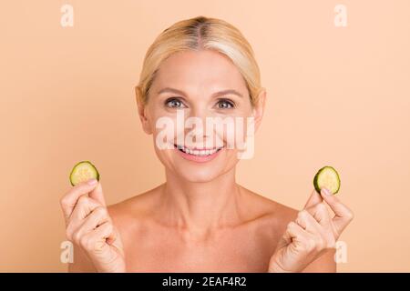 Primo piano ritratto di bionda allegra donna di mezza età che tiene in mano mani cetrioli pezzi isolati su sfondo di colore beige pastello Foto Stock