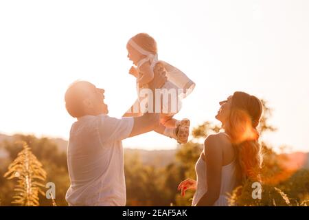 Ritratto di una madre, padre tiene, getta e gira la figlia sulle mani che camminano sulla natura durante la vacanza estiva. Mamma, papà e ragazza che giocano in t Foto Stock