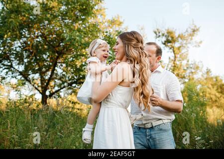 Ritratto di un padre, madre tiene, getta e gira la figlia sulle mani che camminano sulla natura durante la vacanza estiva. Mamma, papà e ragazza che giocano in t Foto Stock