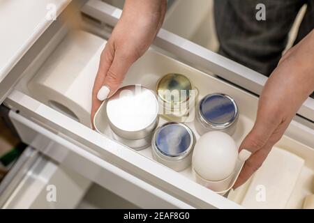 Mani donna che tengono contenitore di plastica con creme e collocano nel cassetto della stanza da bagno. Foto Stock