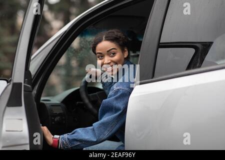 Amichevole sorridente millennial afroamericana driver femminile apre le porte e. cerca il compagno di viaggio Foto Stock