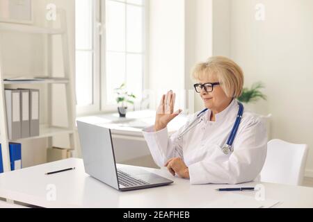 Sorridente anziana professionista donna medico saluto paziente online durante la videocall su un computer portatile Foto Stock