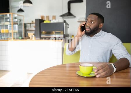 Un ragazzo afro-americano perplesso è seduto nel caffè con una tazza di caffè e ha una conversazione telefonica seria, un uomo multirazziale che parla sullo smartphone Foto Stock